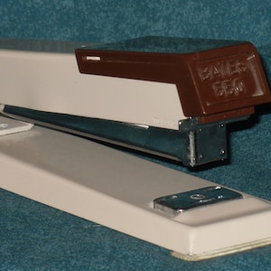 May include: A white and brown stapler with a silver staple remover. The stapler has a brown plastic top with the text "BAKER 556" printed on it.