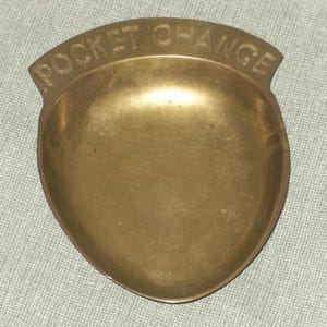 May include: A brass pocket change dish with the words "POCKET CHANGE" embossed across the top. The dish is oval-shaped with a shallow bowl and a slightly raised rim. The brass has a warm, golden hue.