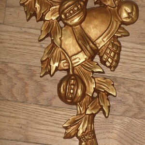 May include: Gold-toned metal wall decor featuring a stylized branch with leaves, fruit, and a bell. The piece has a rustic, vintage aesthetic.