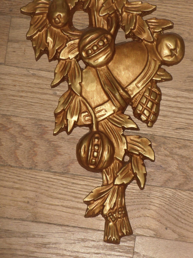 May include: Gold-toned metal wall decor featuring a stylized branch with leaves, fruit, and a bell. The piece has a rustic, vintage aesthetic.