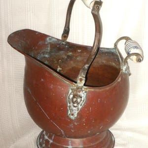 May include: A vintage copper coal scuttle with a rounded body, a flared rim, and a pedestal base. It features a curved handle with white ceramic grips adorned with blue floral patterns. The scuttle shows signs of age and patina.