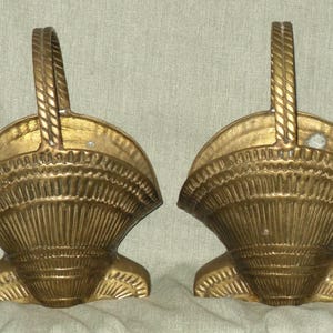 May include: Two gold-colored, decorative basket-shaped objects with handles. The baskets have a fan-like design with vertical lines and a curved handle. The handles are braided. The baskets are made of a metallic material.