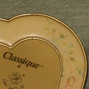 Puede incluir: Un marco de fotos en forma de coraz&oacute;n con un borde dorado y un dise&ntilde;o floral. El marco tiene las palabras "Classique de" y un logotipo en la parte delantera.