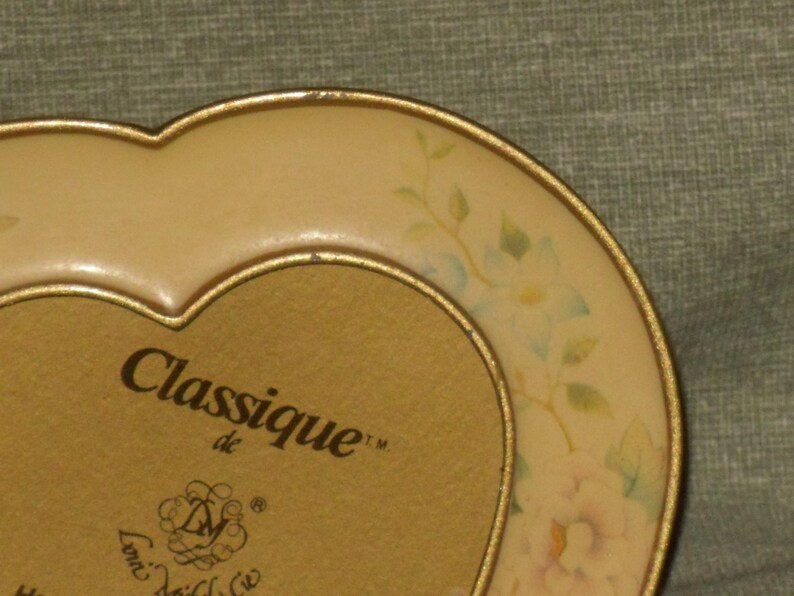 Puede incluir: Un marco de fotos en forma de coraz&oacute;n con un borde dorado y un dise&ntilde;o floral. El marco tiene las palabras "Classique de" y un logotipo en la parte delantera.