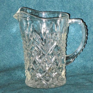 May include: A clear glass pitcher with a diamond pattern. The pitcher has a handle and a spout.