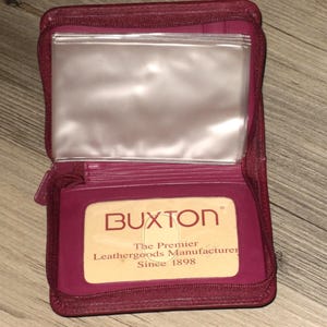 May include: Open burgundy leather wallet with a zipper closure. The interior features clear plastic sleeves and a card holder with the "BUXTON" logo and text: "The Premier Leathergoods Manufacturer Since 1898."