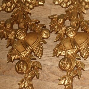 May include: A pair of gold-colored metal wall decor pieces with a floral design and a bell hanging from each. The design features a bow, leaves, and a berry.