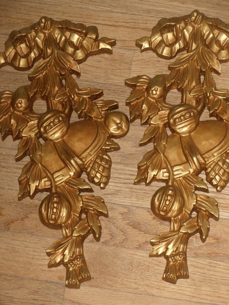 May include: A pair of gold-colored metal wall decor pieces with a floral design and a bell hanging from each. The design features a bow, leaves, and a berry.