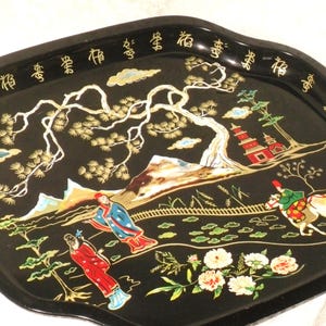 May include: Black decorative tray with a detailed Asian-inspired design. The tray features a landscape with figures, trees, and a pagoda, all in gold and red against a black background. The tray's edge has gold Chinese characters.