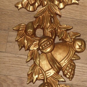 May include: A gold-colored metal ornament featuring a stylized floral design with leaves, berries, and a bell. The ornament has a rustic, vintage aesthetic.