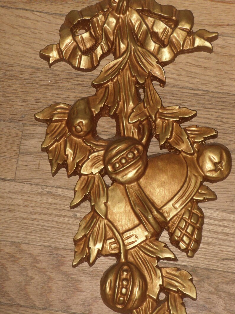 May include: A gold-colored metal ornament featuring a stylized floral design with leaves, berries, and a bell. The ornament has a rustic, vintage aesthetic.