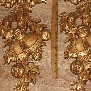 May include: Two gold-painted, ornate, decorative wall hangings with a floral design. The hangings feature a bell, leaves, and a fruit-like design.
