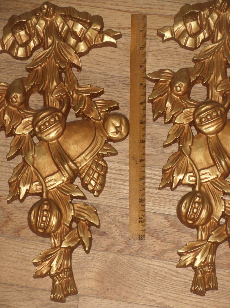 May include: Two gold-painted, ornate, decorative wall hangings with a floral design. The hangings feature a bell, leaves, and a fruit-like design.