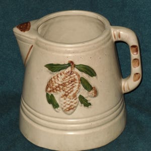 May include: A cream-colored ceramic pitcher with a brown handle and a pine cone design. The pine cone is brown and green with a white background.