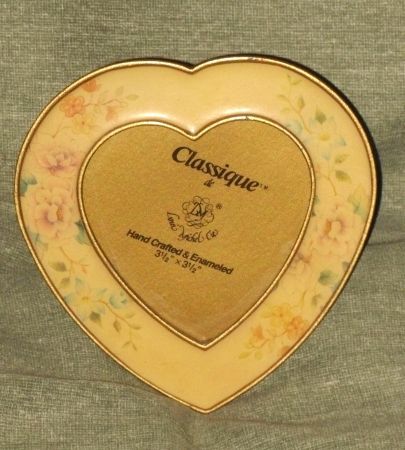 Puede incluir: Un marco de fotos en forma de coraz&oacute;n con un borde dorado y un dise&ntilde;o floral. El marco mide 3 1/2 pulgadas por 3 1/2 pulgadas y dice "Classique de L'Ancienne" y "Hand Crafted & Enameled".