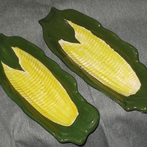 May include: Two ceramic serving dishes shaped like corn on the cob. The dishes are green with a yellow and white corn design. The dishes are on a gray surface.
