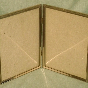 May include: A hinged, double-sided photo frame with a gold-colored metal frame. Each side has a rectangular opening with a cardboard backing. The frame is open, displaying the two sides at an angle.