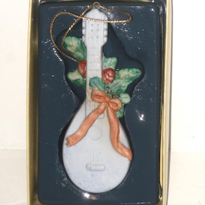 May include: A light blue ceramic mandolin Christmas ornament with a gold string for hanging. The ornament is decorated with green leaves, red berries, and a peach-colored ribbon. The ornament is in a dark blue box with a gold border.