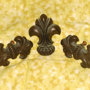 May include: Three ornate, dark brown, metal fleur-de-lis shaped decorative pieces.