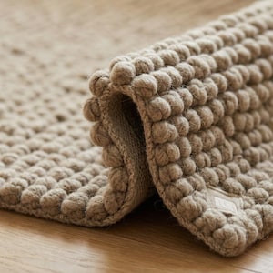 May include: A beige rug featuring a dense, textured surface composed of numerous small, rounded, raised bobbles. The rug is partially folded over, revealing a smooth, flat underside and a small, rectangular fabric label attached to the corner.