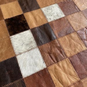 Puede incluir: Una alfombra o tejido de retazos compuesto por cuadrados de cuero y piel cosidos en un patrón de cuadrícula. Los cuadrados presentan diversas texturas y tonos de marrón, bronceado, chocolate oscuro y blanco, incluyendo cuero liso y con pelo.