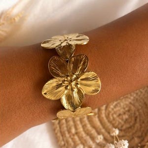 Peut inclure: Un bracelet manchette en métal doré orné de fleurs en relief aux pétales texturés. Le bracelet présente une finition polie et est conçu pour entourer le poignet, mettant en valeur un motif floral répétitif avec des étamines centrales.