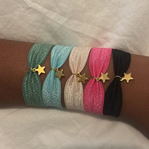 May include: Five elastic hair ties in green, light blue, beige, pink, and black. Each hair tie features a metallic gold star charm attached to the centre. The bands have a shimmering, textured finish and are arranged side by side in a row.