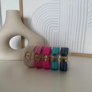 May include: A set of five elastic hair ties arranged in a clear cylindrical container. Each hair tie features a shimmering, textured fabric band in beige, light pink, hot pink, turquoise, and dark blue, accented with a central gold-toned metal H-shaped link.