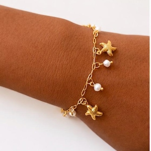 Può includere: Un bracciale a catena color oro con ciondoli alternati a forma di stella marina testurizzata e piccole perle sferiche bianche. Il bracciale è realizzato con maglie ovali e si chiude con un fermaglio, presentando un design nautico.