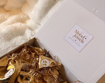 Peut inclure: Une boîte cadeau blanche contenant plusieurs pochettes dorées métallisées à cordon, chacune étiquetée avec un autocollant circulaire blanc portant un numéro. Le couvercle intérieur affiche le texte Mood + Jewels avec un cœur.