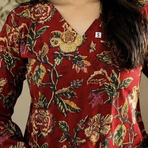 May include: A long-sleeved, V-neck top in a deep red hue, adorned with a floral pattern in shades of yellow, green, and pink. The design features intricate botanical details, creating a rich and textured appearance. A small white tag with the letter "S" is visible.
