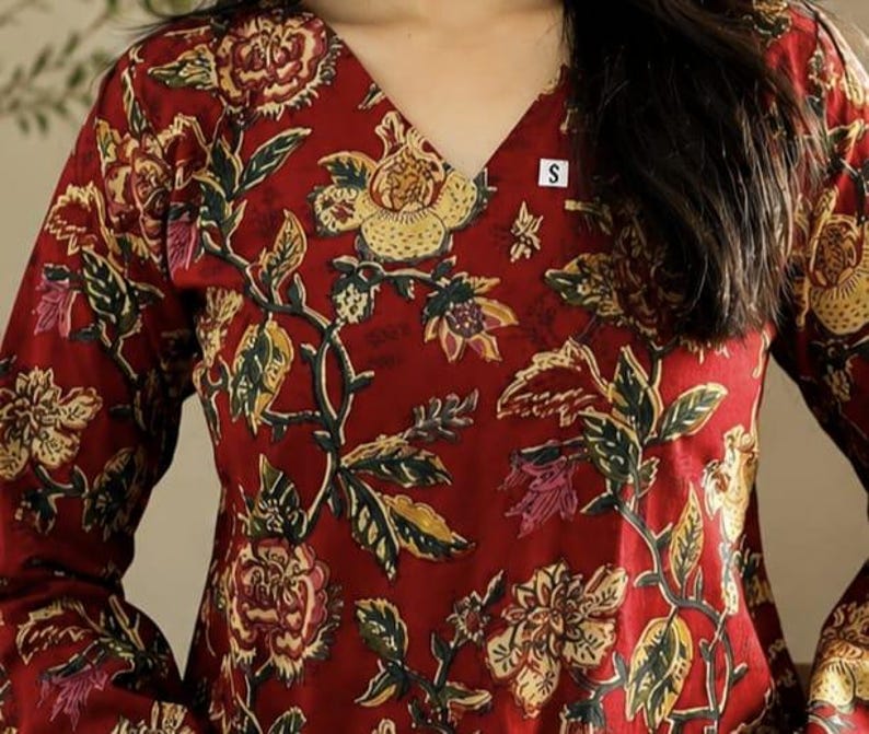 May include: A long-sleeved, V-neck top in a deep red hue, adorned with a floral pattern in shades of yellow, green, and pink. The design features intricate botanical details, creating a rich and textured appearance. A small white tag with the letter "S" is visible.