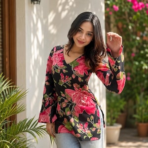 May include: A black tunic top with a V-neck and long sleeves, adorned with a vibrant floral pattern in shades of pink and green. The top is paired with light blue jeans. The background features a white wall, greenery, and a decorative outdoor light.