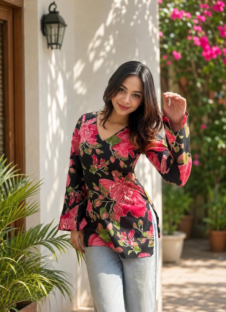 May include: A black tunic top with a V-neck and long sleeves, adorned with a vibrant floral pattern in shades of pink and green. The top is paired with light blue jeans. The background features a white wall, greenery, and a decorative outdoor light.