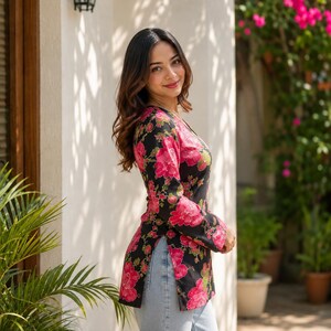 May include: A black top with a vibrant pink floral pattern. The long-sleeved top has a V-neck and side slits, paired with light blue jeans. The image is taken outdoors, with a white wall and greenery in the background.