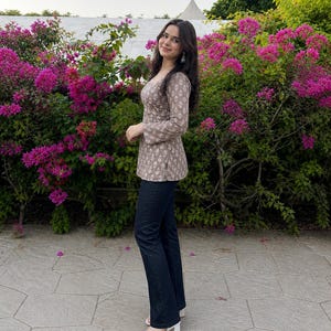 Puede incluir: Una mujer con una blusa de manga larga color topo con estampado floral blanco y vaqueros azul oscuro. Lleva sandalias blancas de tac&oacute;n. El fondo presenta flores rosas vibrantes y follaje verde.