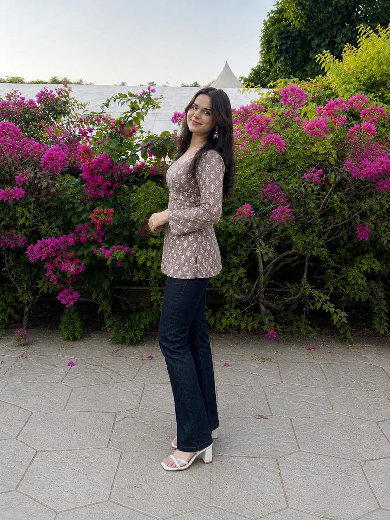 Puede incluir: Una mujer con una blusa de manga larga color topo con estampado floral blanco y vaqueros azul oscuro. Lleva sandalias blancas de tac&oacute;n. El fondo presenta flores rosas vibrantes y follaje verde.