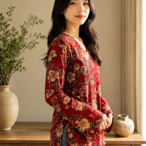 May include: A long-sleeved, red blouse with a V-neck and a floral pattern in yellow and green. The blouse is paired with blue jeans. The image is taken indoors, with a wooden table and a vase in the background.