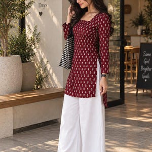 May include: A long-sleeved maroon tunic featuring a white floral print, a square neckline, and side slits, paired with loose-fitting white wide-leg trousers and white strappy sandals. A black and white houndstooth patterned tote bag is carried.