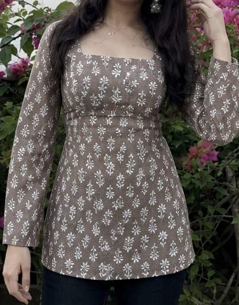 Puede incluir: Una blusa marr&oacute;n de manga larga con cuello cuadrado y estampado floral blanco. La blusa tiene cintura entallada y bajo ligeramente acampanado. Las mangas son largas y ajustadas. El fondo presenta flores rosas y follaje verde.