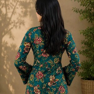 May include: A teal long-sleeve top with a floral pattern in red, yellow, and green. The top has a fitted silhouette and flared sleeves. The model is wearing blue jeans. The background is a neutral color.