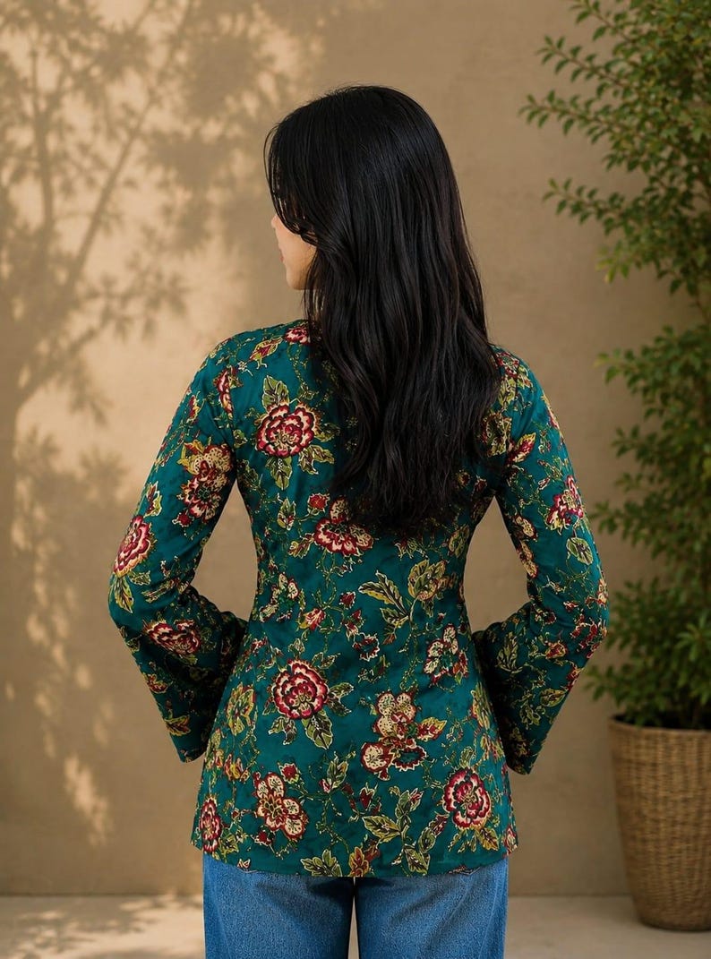 May include: A teal long-sleeve top with a floral pattern in red, yellow, and green. The top has a fitted silhouette and flared sleeves. The model is wearing blue jeans. The background is a neutral color.