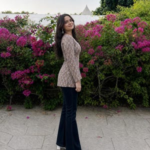 Puede incluir: Una mujer con un top marr&oacute;n con estampado floral y pantalones acampanados azul oscuro. Tambi&eacute;n lleva sandalias blancas. El fondo presenta flores rosas vibrantes y follaje verde.