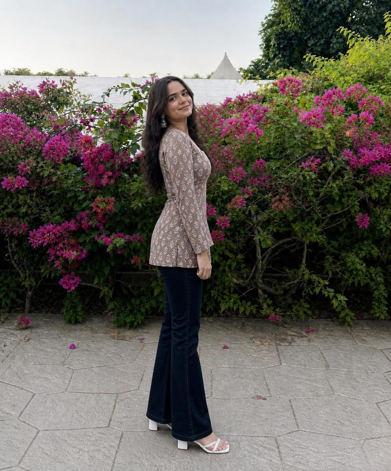 Puede incluir: Una mujer con un top marr&oacute;n con estampado floral y pantalones acampanados azul oscuro. Tambi&eacute;n lleva sandalias blancas. El fondo presenta flores rosas vibrantes y follaje verde.