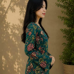 May include: A woman wearing a teal floral print top with long sleeves and a V-neck. The top features a pattern of red and yellow flowers and green leaves. She is wearing blue jeans. A potted plant is in the background.