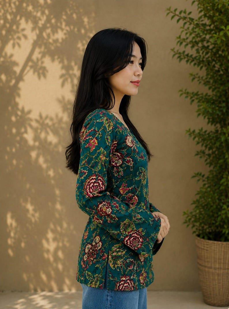 May include: A woman wearing a teal floral print top with long sleeves and a V-neck. The top features a pattern of red and yellow flowers and green leaves. She is wearing blue jeans. A potted plant is in the background.