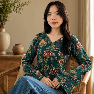 May include: A long-sleeved, teal blouse with a V-neck and a floral pattern in red, yellow, and brown. The blouse is paired with blue jeans. The model is seated in a wooden chair.