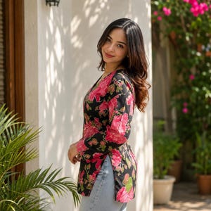 May include: A black floral print top with pink flowers and green leaves. The top has long sleeves and a V-neck. The person is wearing light blue jeans. The background includes a white wall, plants, and a black outdoor light fixture.