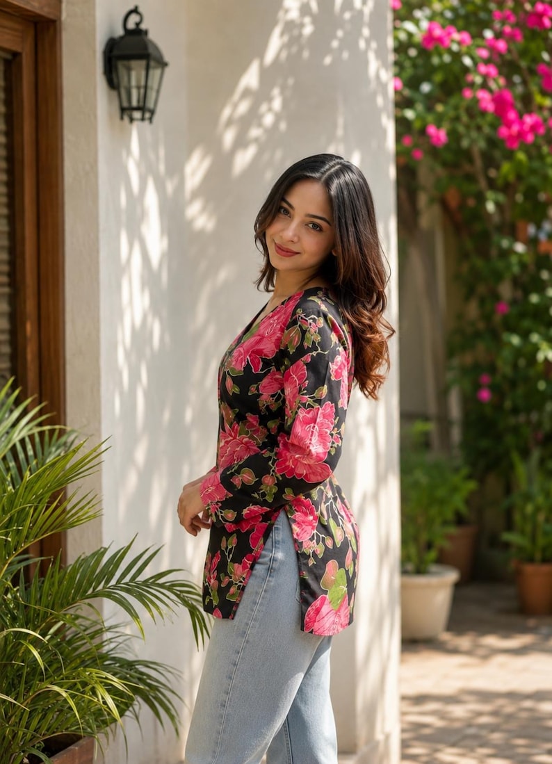 May include: A black floral print top with pink flowers and green leaves. The top has long sleeves and a V-neck. The person is wearing light blue jeans. The background includes a white wall, plants, and a black outdoor light fixture.