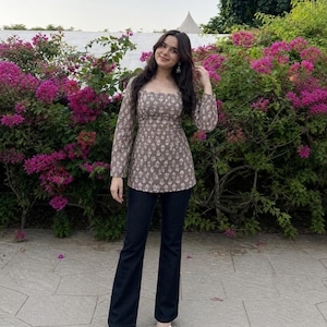 May include: A brown floral print top with long sleeves and a square neckline, worn with dark wash flared jeans. The outfit is completed with white sandals. The background features vibrant pink flowers and green foliage.